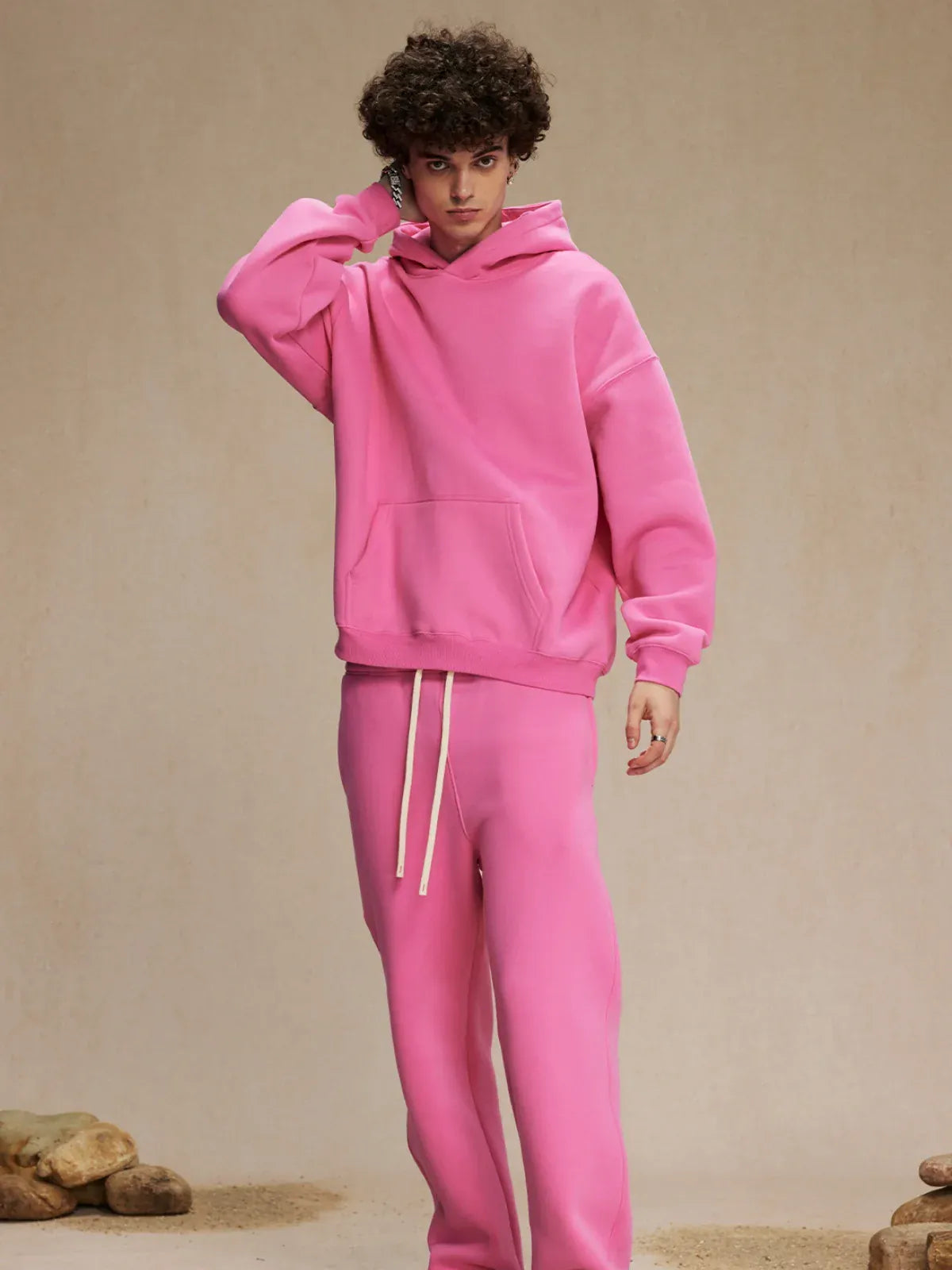 A person stands indoors in the Custom Anime Character Bubblegum Pink Hoodie - Personalized 350GSM 100% Cotton Pullover and matching sweatpants, touching the back of their neck. Rocks are visible on the floor by their feet.