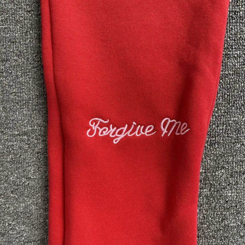 The Syna World Red Script Embroidered Drawstring Jogger Pants feature "Forgive Me" in white cursive on red fabric, styled atop a gray textured background.