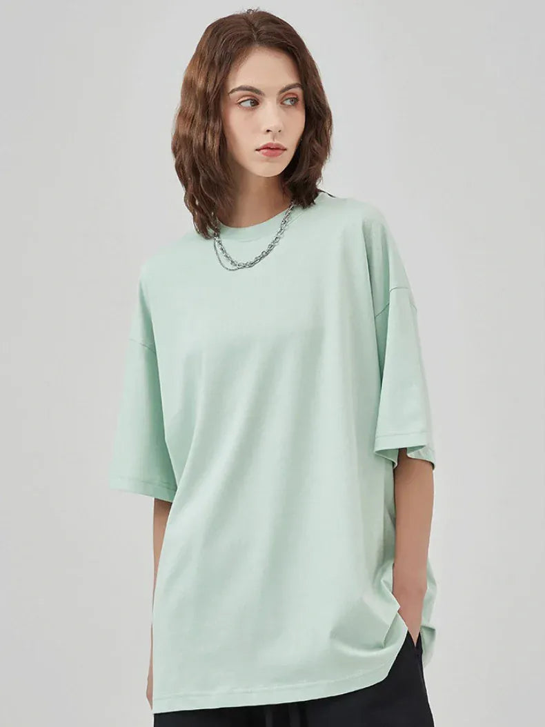 A woman with shoulder-length brown hair wears the Custom Anime Character Mint Green T-Shirt, made from 230GSM 100% cotton, paired with black pants and a silver chain necklace, standing against a plain light gray background.