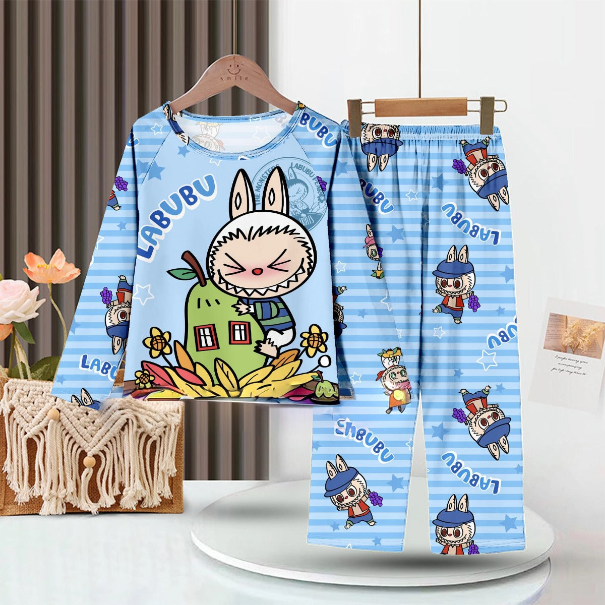 Displayed on a hanger, the LABUBU Baby Pajama Set features a large-toothed, bunny-eared cartoon on blue stripes. Crafted from breathable milk silk fabric, &quot;Labubu&quot; is printed on both top and pants, surrounded by floral designs and a woven bag.