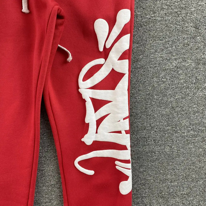 Syna World Red Letter Print Drawstring Jogger Pants feature bold white graffiti-style text along the leg, an abstract print with curved lines and dots on a gray textured surface, and are crafted from soft cotton-polyester joggers material.