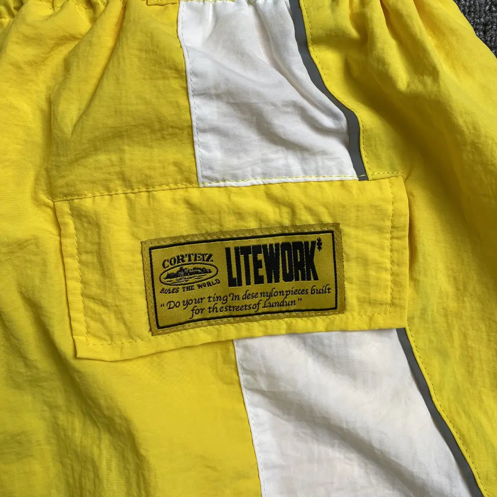 Close-up of Corteiz Yellow & White Colorblock Nylon Shorts, featuring a white vertical stripe, elastic waistband, and a stitched patch: “Corteiz LITEWORK* ‘Do your thing. In destiny pieces built for the streets of Landan’.”.
