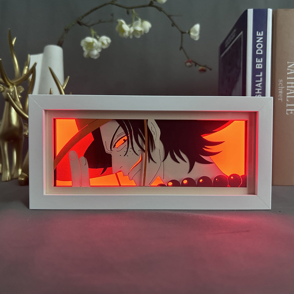 The Seakoff One Piece Anime Light Box features a 3D LED lamp of Portgas D. Ace from One Piece with color-changing effects, including orange-red. It adorns a table, complemented by a book and deer head decor with white flowers above.