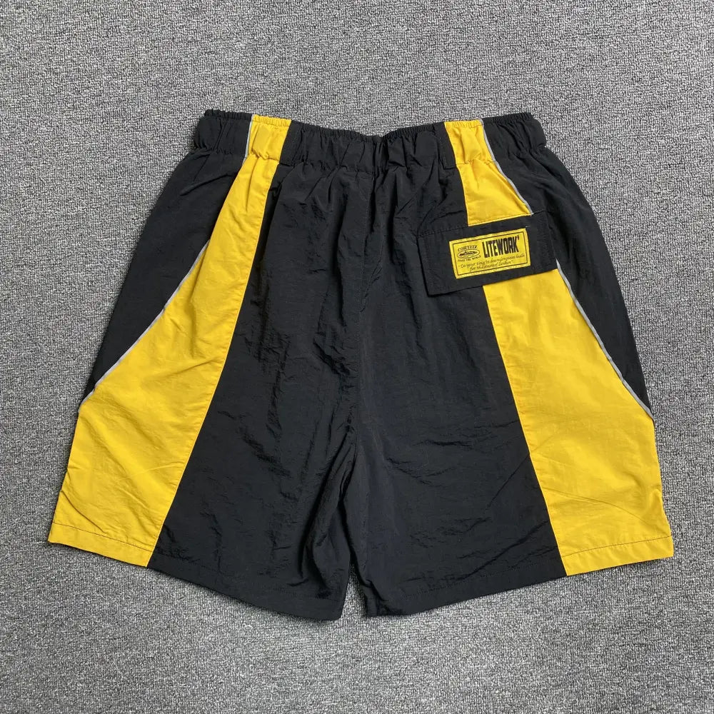 Corteiz Black Yellow Colorblock Nylon Shorts with a relaxed fit, featuring black and bright yellow panels and a yellow text patch on the back, shown laid flat on a gray carpeted surface.