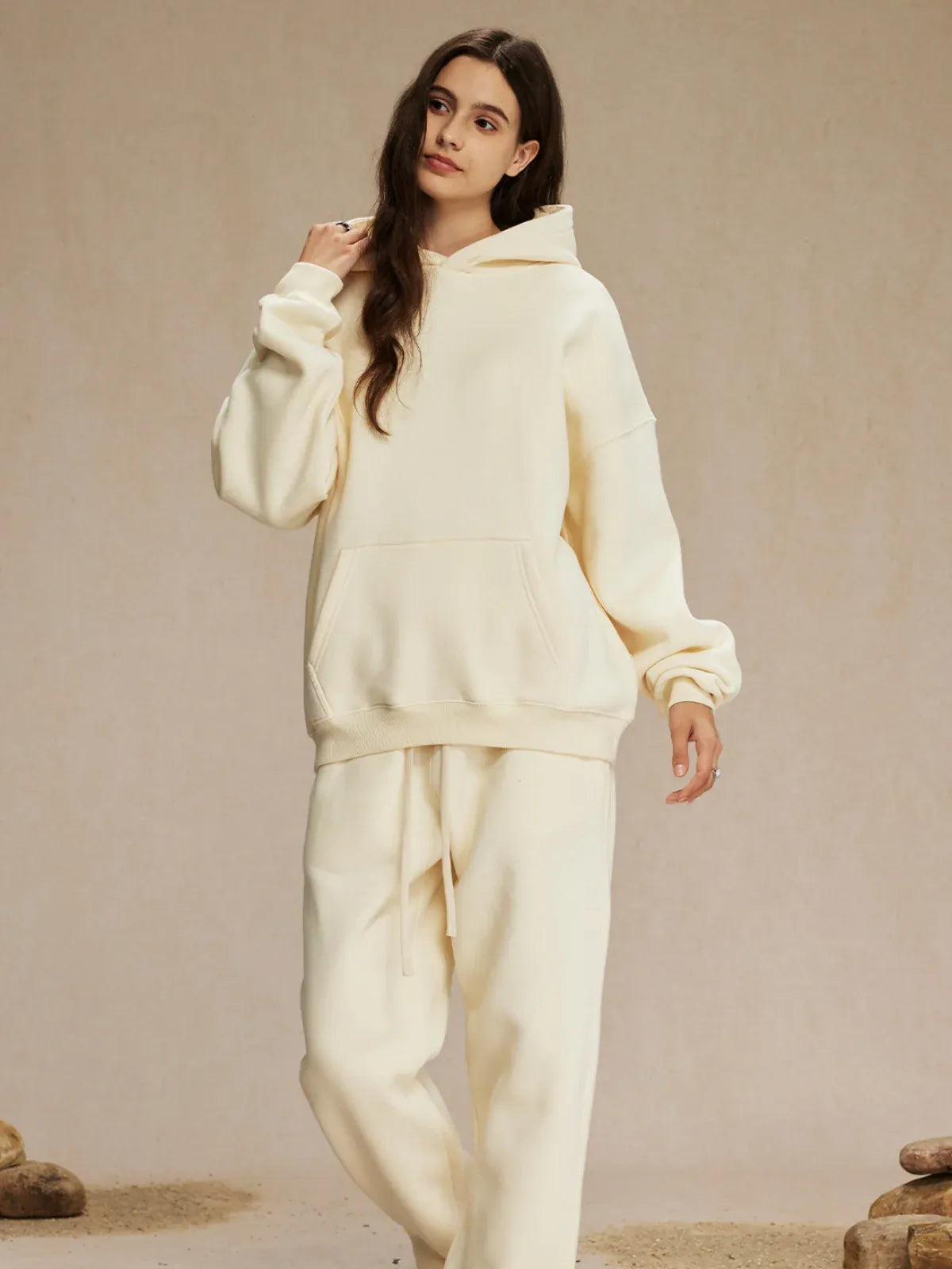 A woman stands indoors wearing a Custom Anime Character Cream Hoodie – Personalized 350GSM 100% Cotton Pullover, paired with matching sweatpants, her left hand raised near her head.