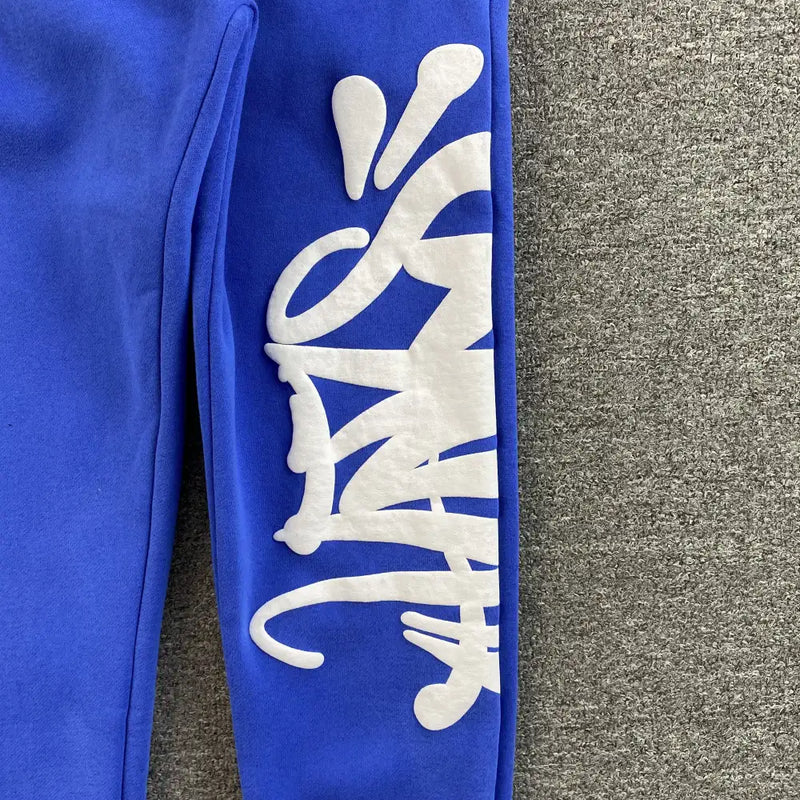 The Syna World Blue Graffiti Graphic Jogger Pants feature a royal blue sleeve with bold white graffiti-style vertical lettering, set against a textured gray background.