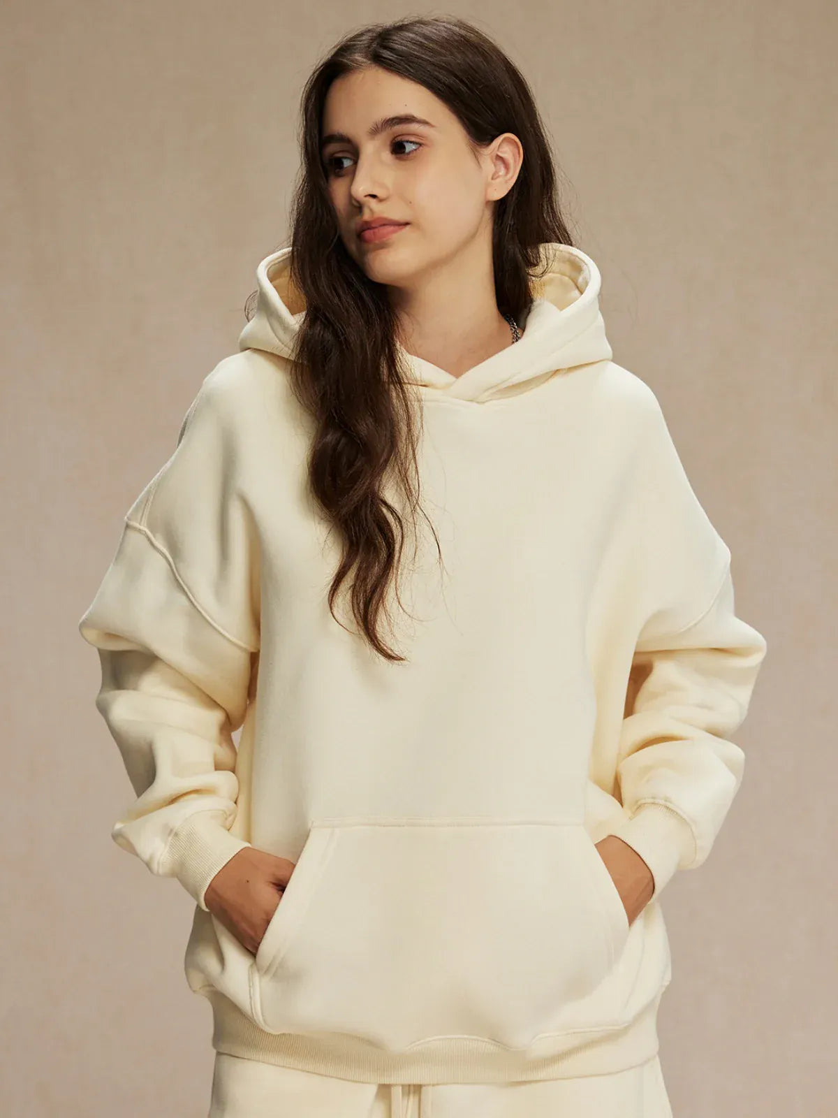 A person with long brown hair stands with hands in the front pocket, wearing the Custom Anime Character Cream Hoodie - Personalized 350GSM 100% Cotton Pullover and looking slightly to the side.