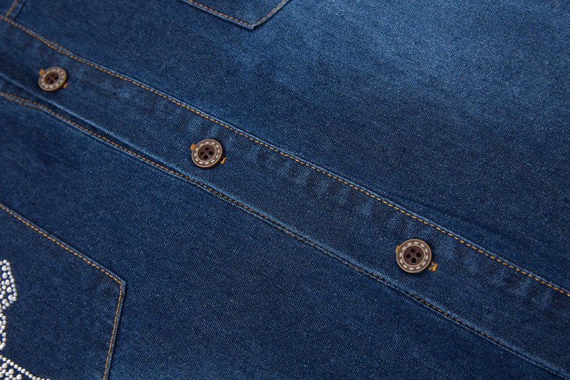 Close-up of a blue denim button-up shirt with tan stitching, chest pocket trim, and rhinestone cross detailing