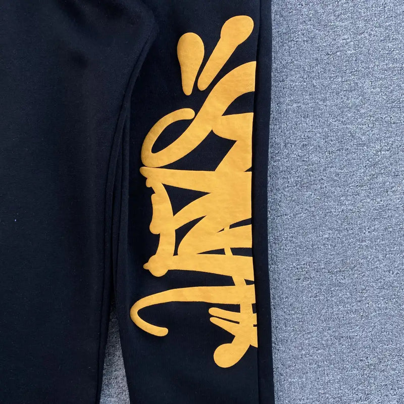 The Syna World Black Yellow Graffiti Print Jogger Pants feature bold, yellow graffiti-style text on black fabric, offering an urban edge—perfect for pairing with your favorite streetwear outfits.
