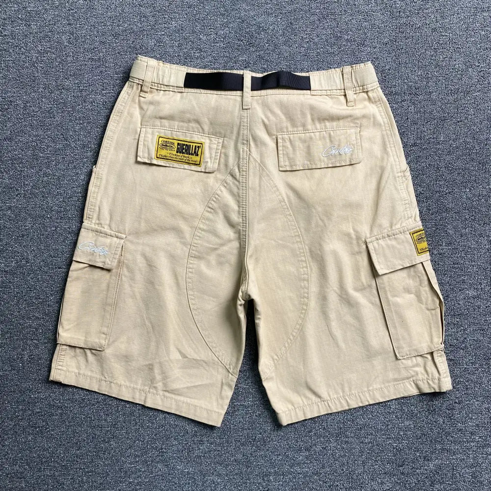 Corteiz Beige Cargo Shorts with front graphic, featuring multiple pockets, black belt loops for a webbing belt, and yellow rectangular patches, displayed flat on a gray textured surface.