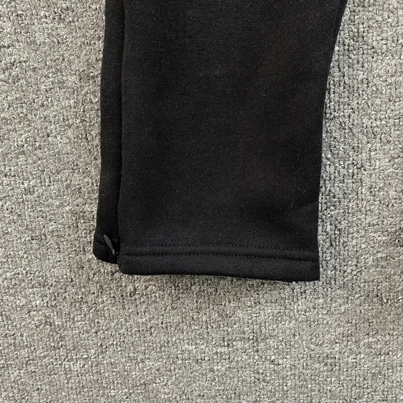Close-up of the hem of Syna World Black Yellow Graffiti Print Jogger Pants on a gray textured carpet. The smooth fabric, slightly folded at the seam, highlights their modern streetwear style.