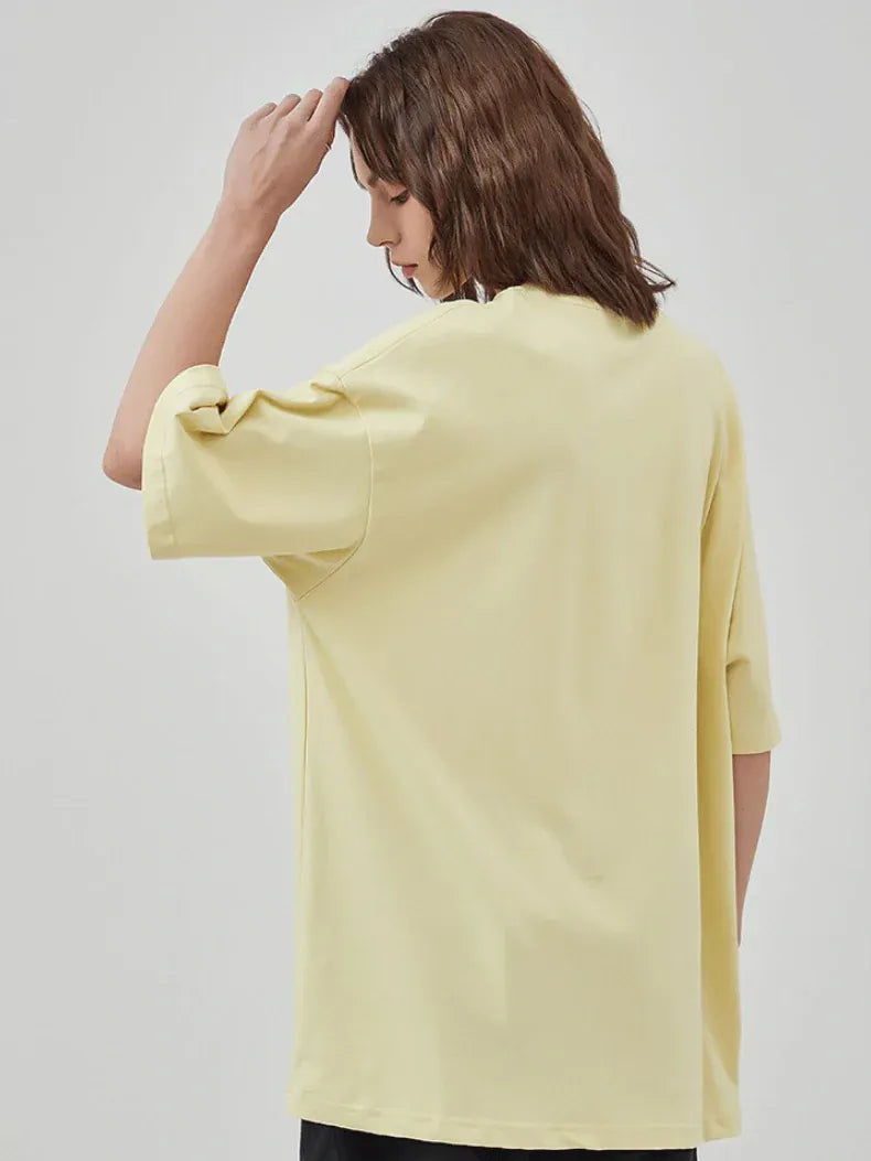 A person with shoulder-length brown hair wears the Custom Anime Character Butter Yellow T-Shirt, a loose-fit 100% cotton 230GSM tee, standing with their back to the camera and touching their head.