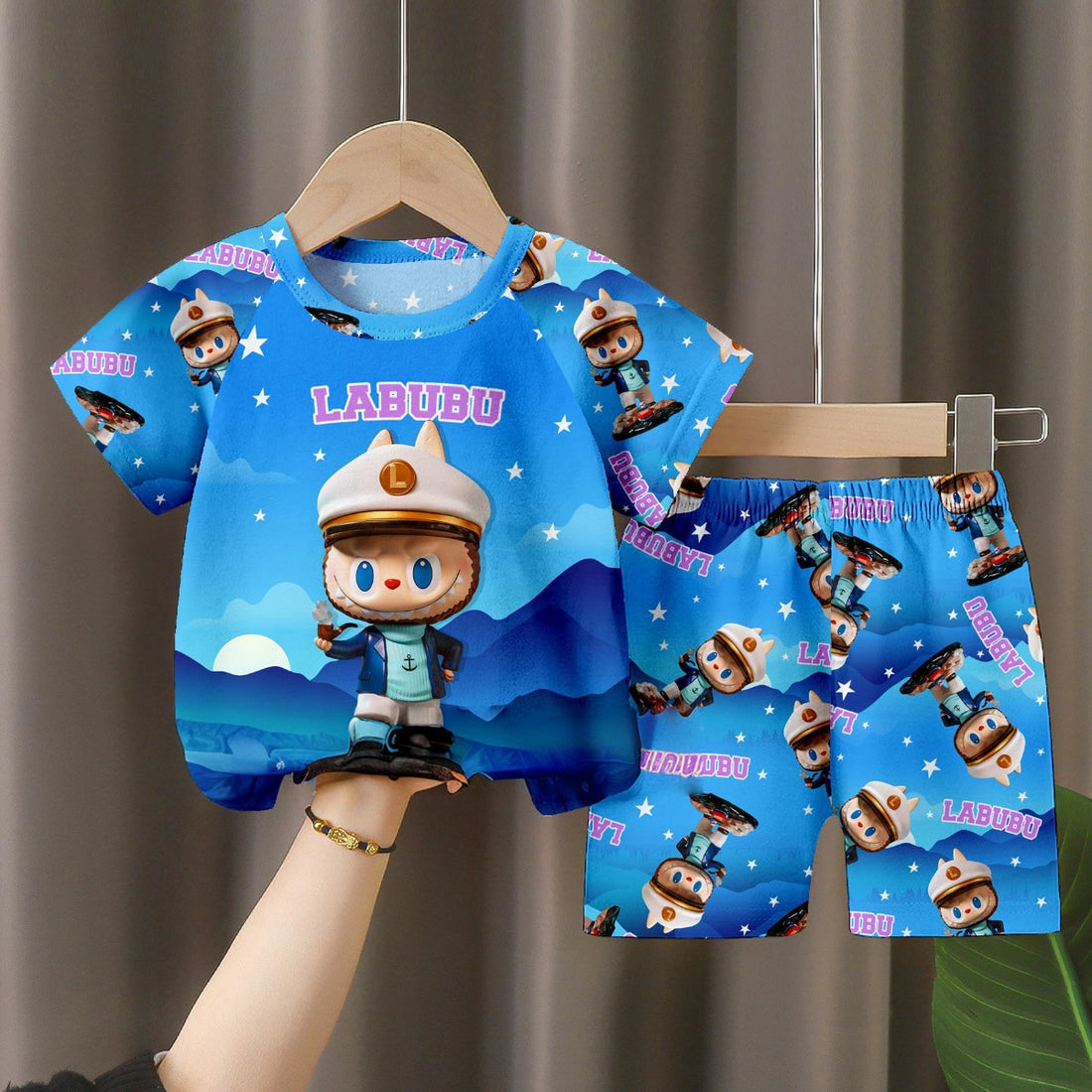 A hand with a bracelet displays the LABUBU Baby Pajama Set, featuring a blue T-shirt and shorts made from soft milk silk. Both pieces spotlight the LABUBU character with large eyes, a hat, an outfit, and a starry backdrop with &quot;Labubu&quot; repeated.