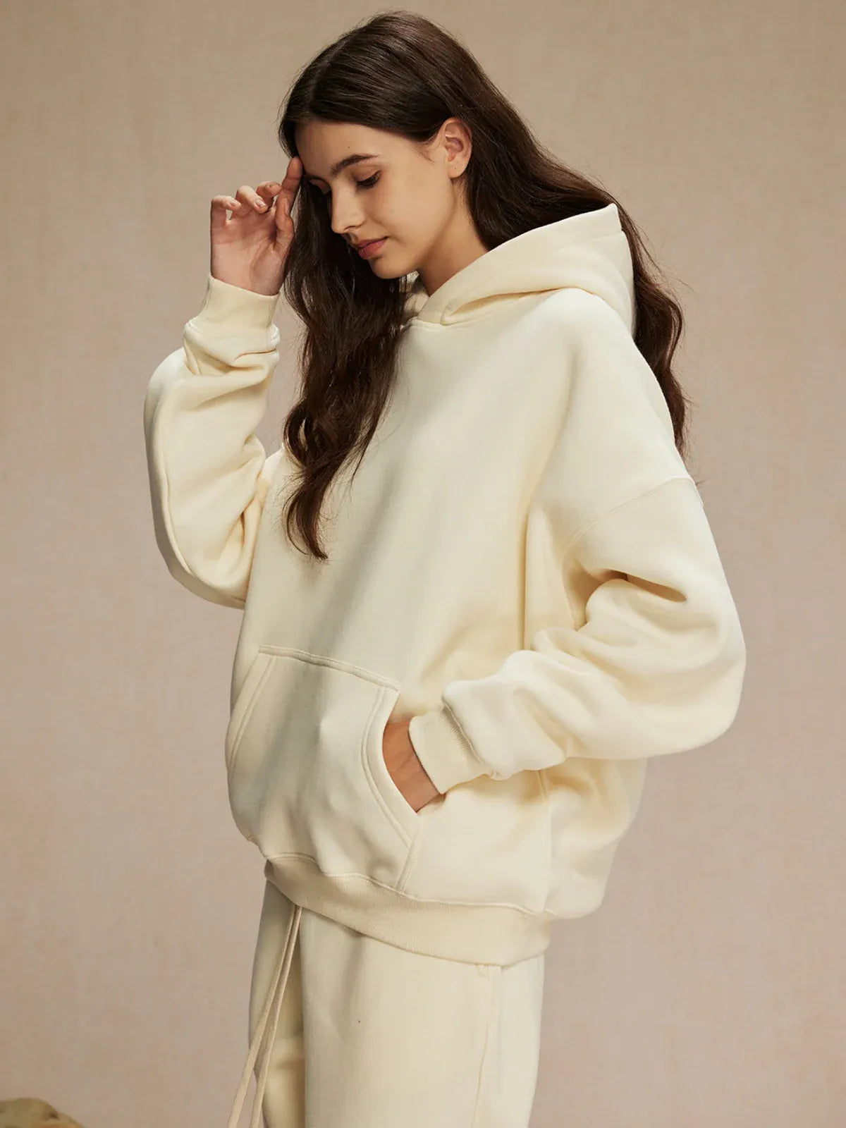 A person with long brown hair wears the Custom Anime Character Cream Hoodie—Personalized 350GSM 100% Cotton Pullover—with matching sweatpants, standing with one hand in their pocket and the other touching their head.