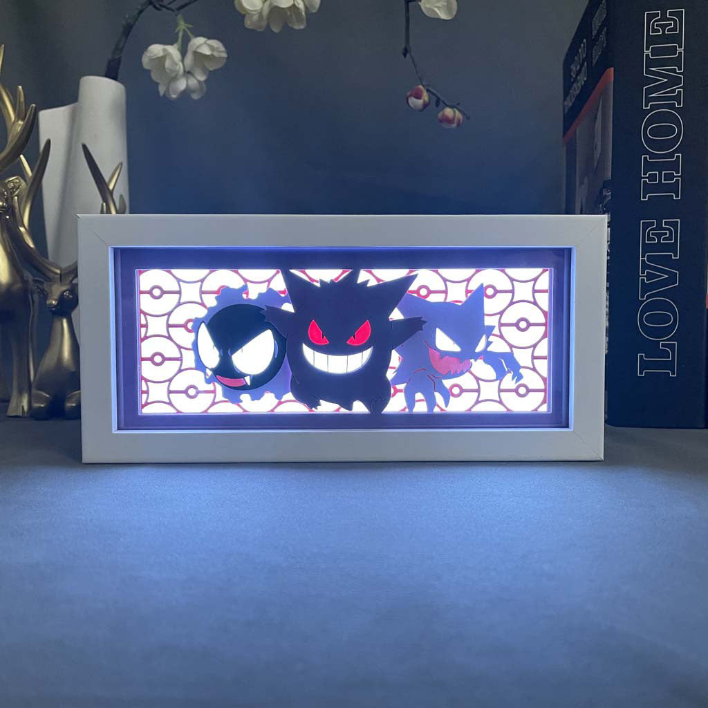 The Pokémon Gastly, Haunter & Gengar 3D LED Light Box highlights a black figure with red eyes against an abstract backdrop. The glowing frame, adorned with decorative elements like flowers and a statue, showcases the Ghost Trio reminiscent of Pokémon fan favorites.