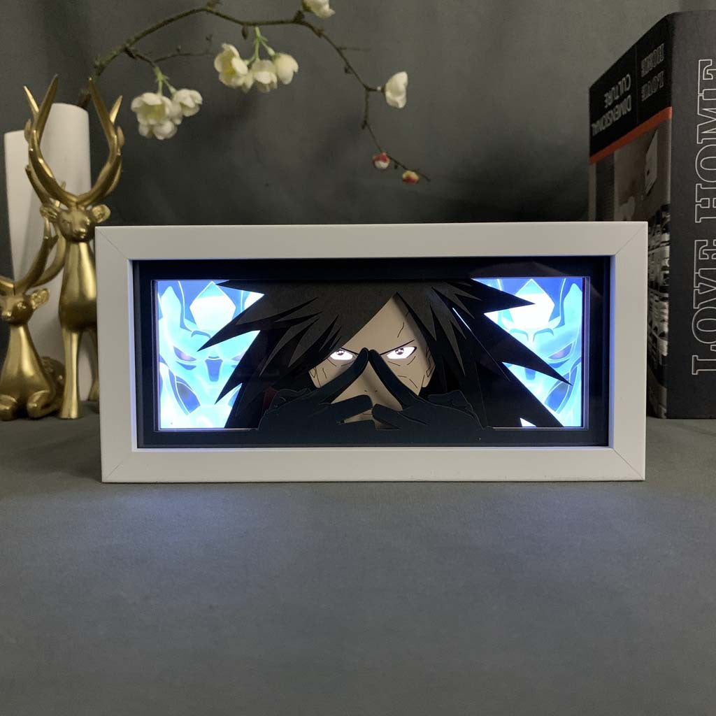 The Naruto Madara Uchiha LED 3D Light Box – Susanoo Warrior Design, featuring Madara with long dark hair against a blue abstract background, glows on a shelf surrounded by decorative items like a golden reindeer and a book.