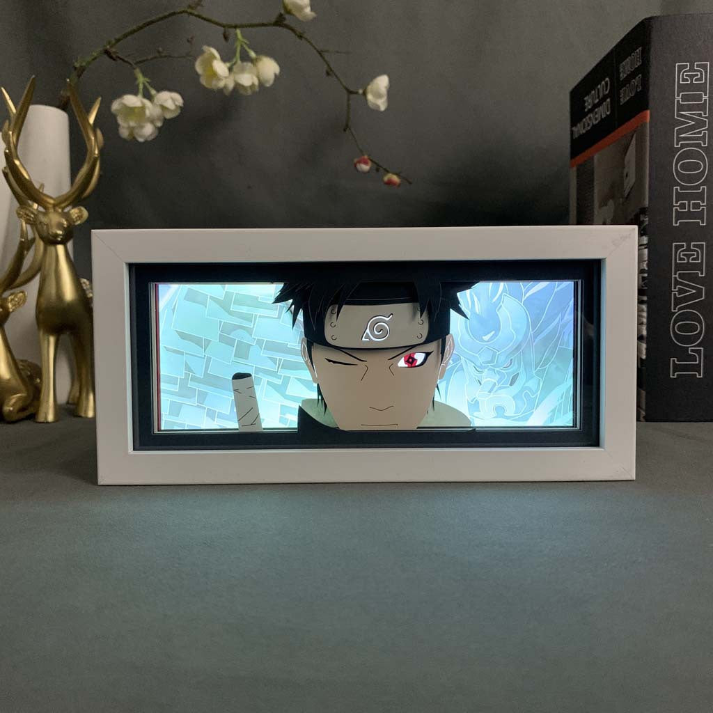 The Uchiha Shisui LED Anime Light Box – Naruto 3D Night Light Decor by Naruto displays an anime character with a headband and red eye on the desk. Nearby, a gold deer figure and books, including &quot;LOVE HOME&quot; with white flowers, enhance the scene. Perfect for Naruto fans!.