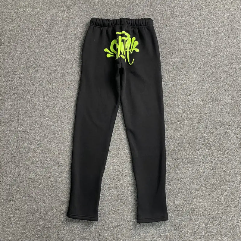 The Syna World Black Letter Print Drawstring Jogger Pants feature an elastic waistband and neon green lettering near the waist—an ideal choice for bold streetwear style, shown laid flat on a gray carpeted surface.