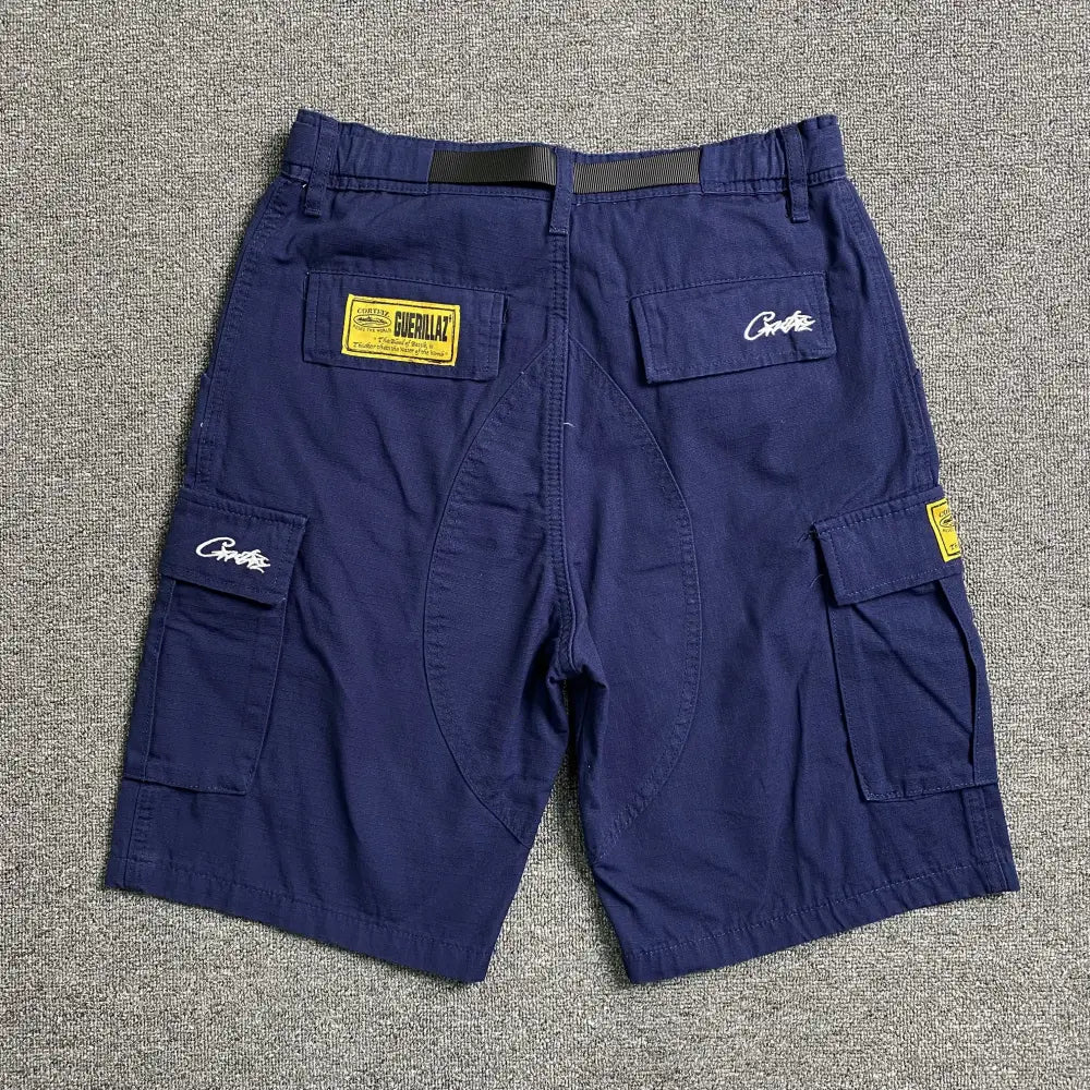 Corteiz Blue Cargo Shorts with Front Graphic Print, featuring multiple flap pockets and bold logos, showcased on a gray carpet—ideal streetwear shorts for your collection.