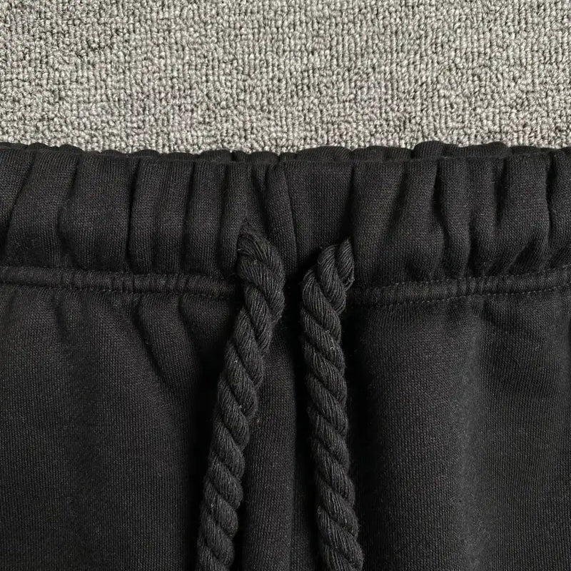 Close-up of the waistband on Syna World Black Letter Print Drawstring Jogger Pants, featuring thick twisted drawstrings and neon green lettering, shown on a textured light gray carpet.