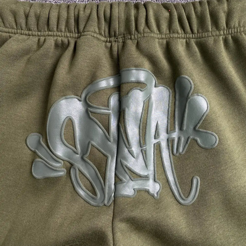 Close-up of Syna World Olive Green Drawstring Jogger Pants in a cotton-polyester fleece, featuring a raised glossy gray graffiti-style design with abstract overlapping letters and shapes.