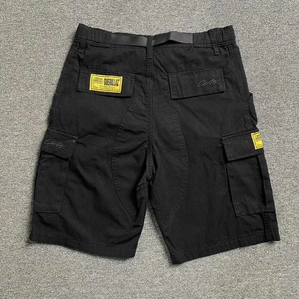 The Corteiz Black Cargo Shorts with Front Graphic feature multiple pockets, yellow rectangular patches, and black embroidered text on the back—ideal for any streetwear shorts collection. Displayed flat on a grey textured surface.