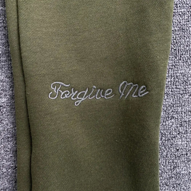 Syna World Olive Green Drawstring Jogger Pants, made from cotton polyester fleece with "Forgive Me" in light gray cursive embroidery, displayed on a textured gray surface.