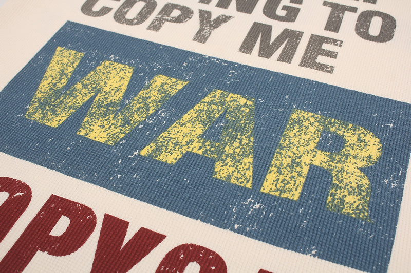 Close-up of a waffle knit tee with distressed WAR graphic in yellow on blue and partial COPY TO ME text on white fabric