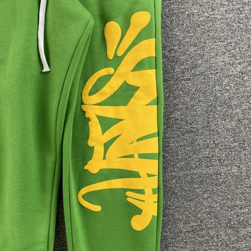 Syna World Green Yellow Letter Print Drawstring Jogger Pants feature a white drawstring and bold yellow graffiti-style text on the left leg, styled as streetwear against a gray textured background.