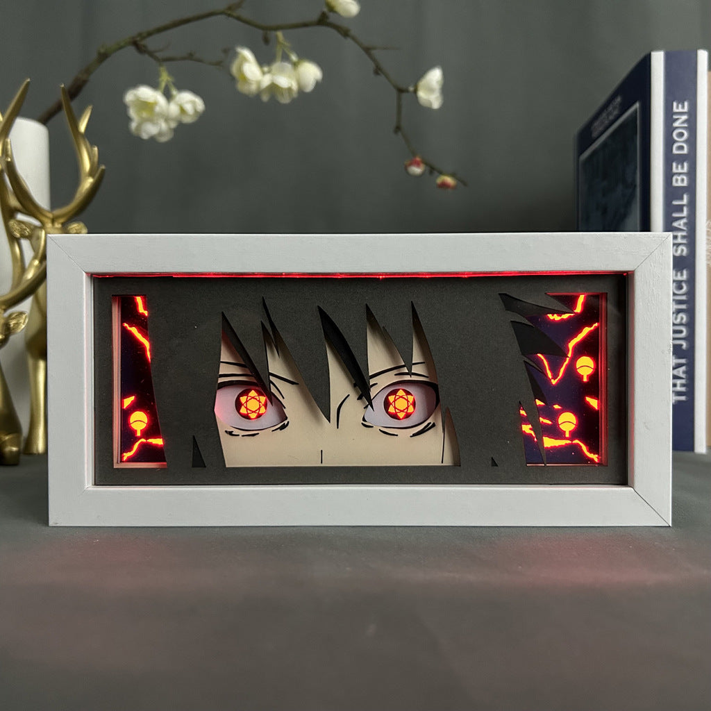 This Naruto Anime Light Box showcases a 3D effect with stylized eyes reminiscent of the Mangekyou Sharingan against a glowing red backdrop. Featuring a 16-color adjustable LED light, this Sasuke Uchiha frame sits on a surface with decorative branches and a book, making it an intriguing collectible.