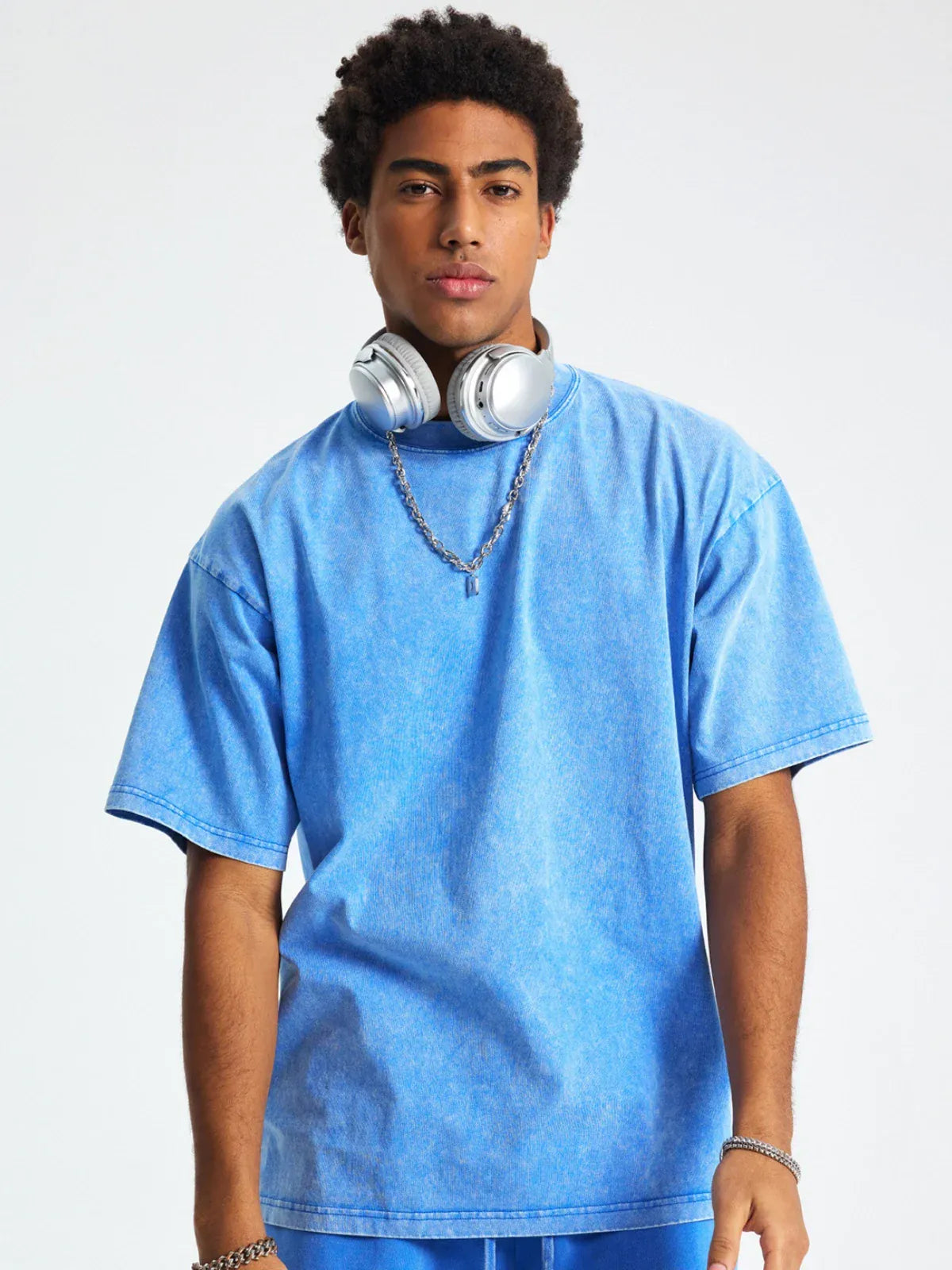 A young man with short curly hair wears the Custom Anime Character Blue Washed T-Shirt, paired with a silver chain necklace and headphones around his neck, standing against a plain light background.