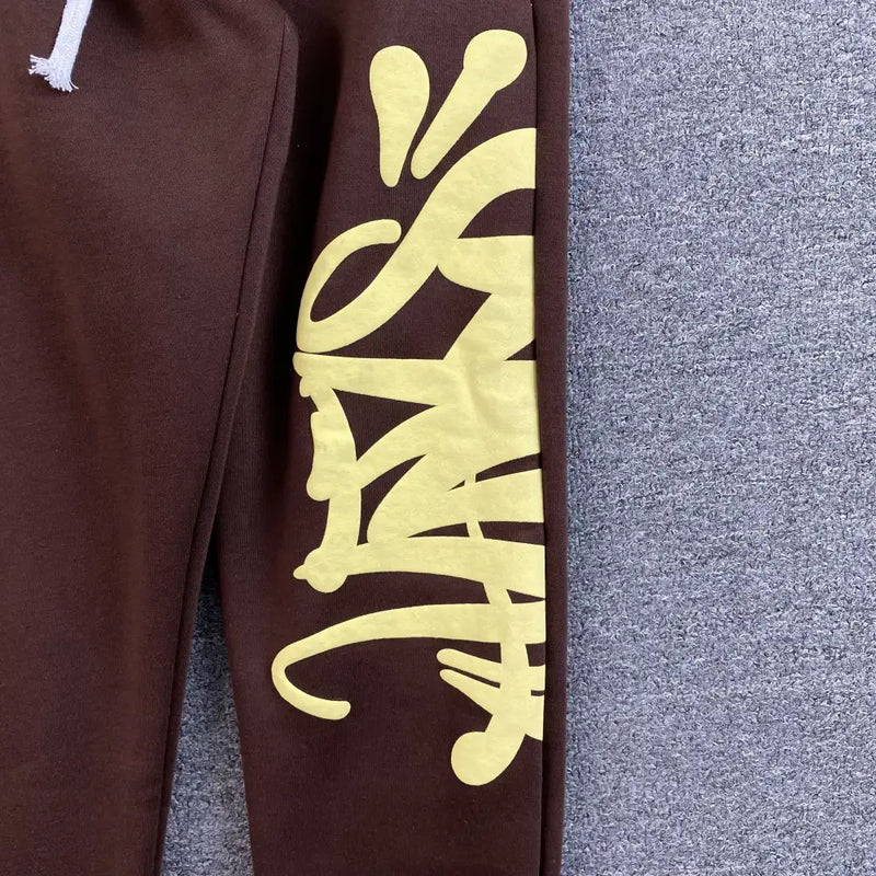 Syna World Brown Yellow Graffiti Print Joggers feature bold yellow graffiti lettering running vertically along one leg, lending a streetwear vibe. Only a portion of these statement joggers is displayed on a gray textured surface.
