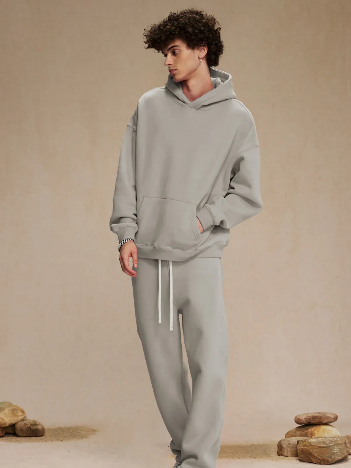 Indoors, a person wears the Custom Anime Character Light Gray Hoodie—Personalized 350GSM 100% Cotton Pullover. The oversized hoodie pairs with matching sweatpants; one hand is in their pocket while small rocks are scattered on the floor.