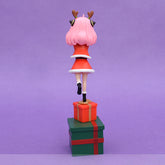 The Anya Forger Christmas PVC Figure - 18cm Holiday Edition by Spy × Family features pink-haired Anya in a red dress with white trim and black shoes, standing on tiptoe atop a red gift box with an antler headband. Below is a larger green gift box, all set against a plain purple background.