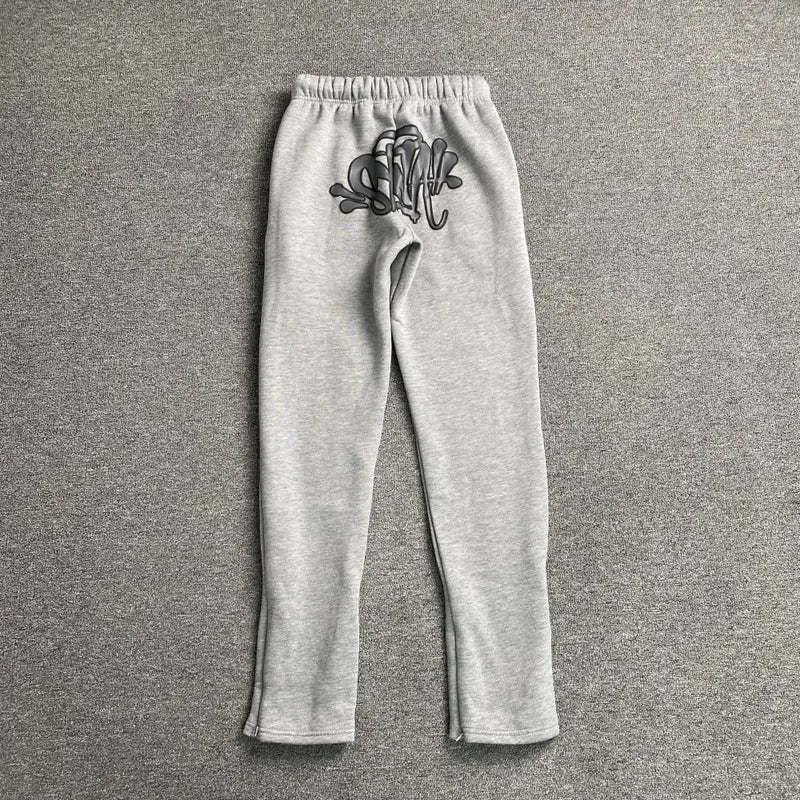 The Syna World Light Grey Letter Print Drawstring Jogger Pants, made from a cotton-polyester blend with an elastic waistband and black graffiti-style print on the left hip, are displayed flat on a matching gray textured surface.