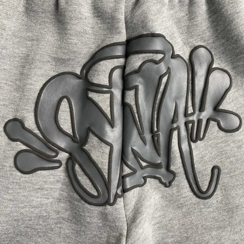 Close-up of Syna World Light Grey Letter Print Drawstring Jogger Pants in a cotton-polyester blend, featuring black graffiti-style "SNAK" lettering outlined in raised, shiny material with decorative splash accents.