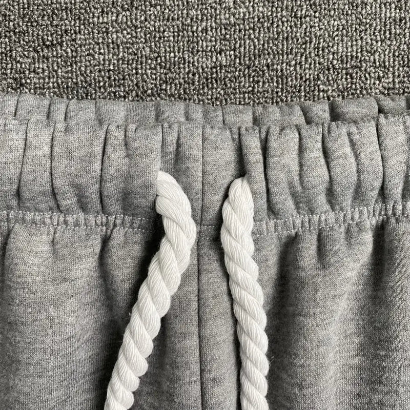 Close-up of the elasticated waistband and thick, twisted white drawstrings on Syna World Light Grey Letter Print Drawstring Jogger Pants, shown against a textured gray fabric background.