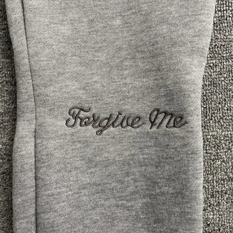 Close-up of Syna World Light Grey Letter Print Drawstring Jogger Pants in a soft cotton-poly blend, featuring an elasticated drawstring waist and "Forgive Me" embroidered in dark gray cursive on textured fabric.