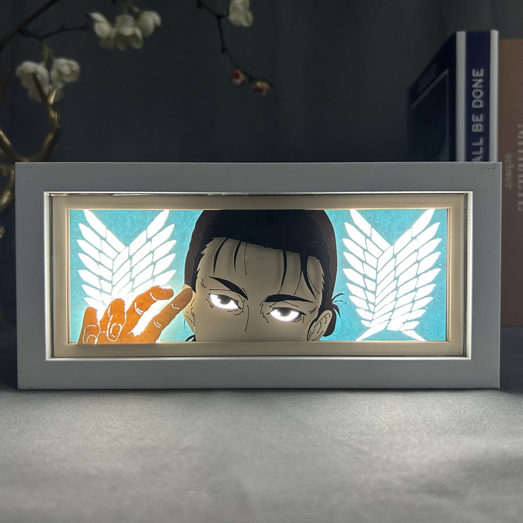 The Attack on Titan Anime Light Box showcases a 3D effect of Eren Yeager with short dark hair and intense eyes. Set in a rectangular frame, it features his character against a blue backdrop with wing emblems, plus partial views of a book and flowers. It offers 16-color adjustable LED light settings.