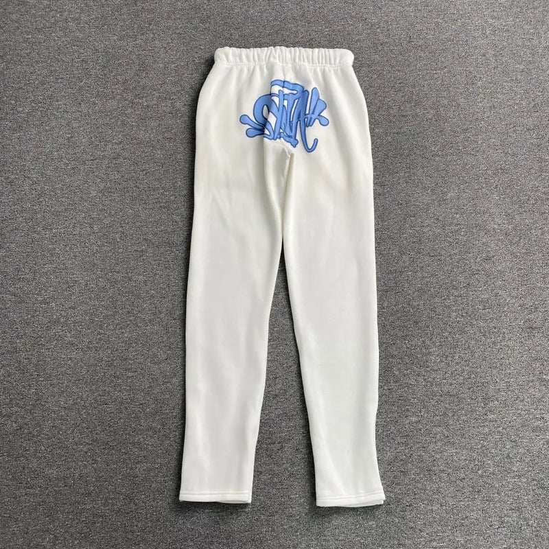 Syna World White Forgive Me Script Jogger Pants feature an elastic waistband and bold blue graffiti-style text on the upper back. Made from a cotton-polyester blend, they are displayed lying flat on a gray surface.