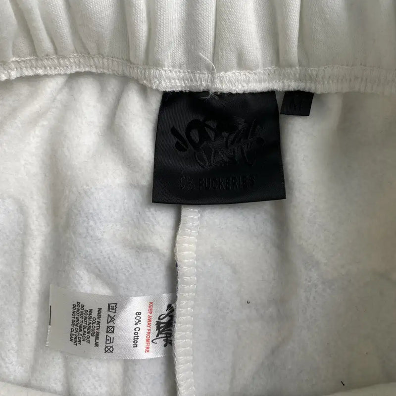 Close-up of the inside waistband of Syna World White Forgive Me Script Jogger Pants, featuring two black and white tags: one with O2: FUCKERIES branding and the other with care instructions and 80% cotton material details.