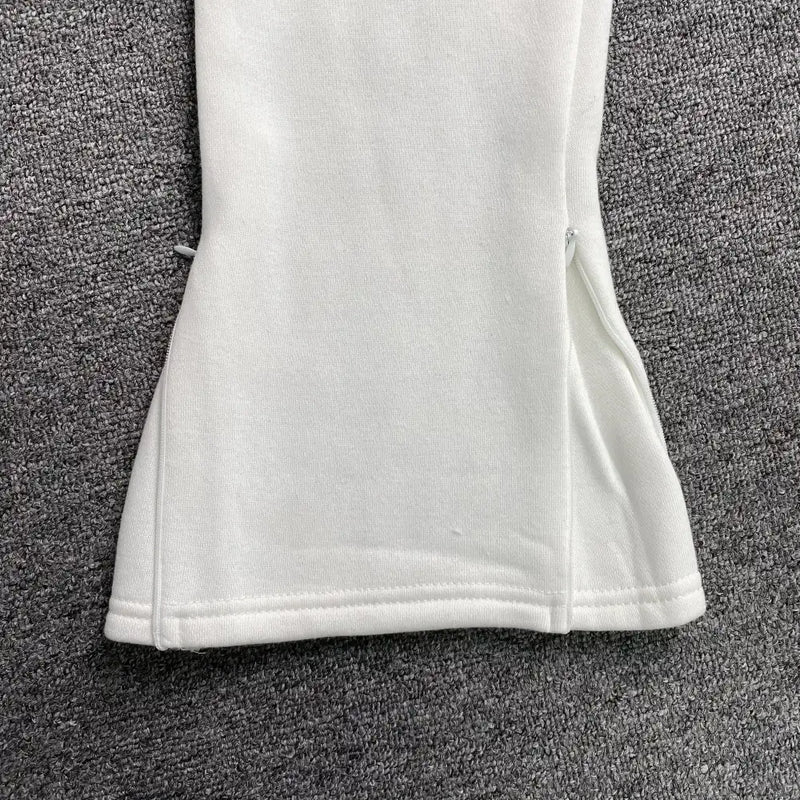 Close-up of the lower part of Syna World White Forgive Me Script Jogger Pants, showing a short concealed ankle zip near the hem, displayed on a gray textured carpet.