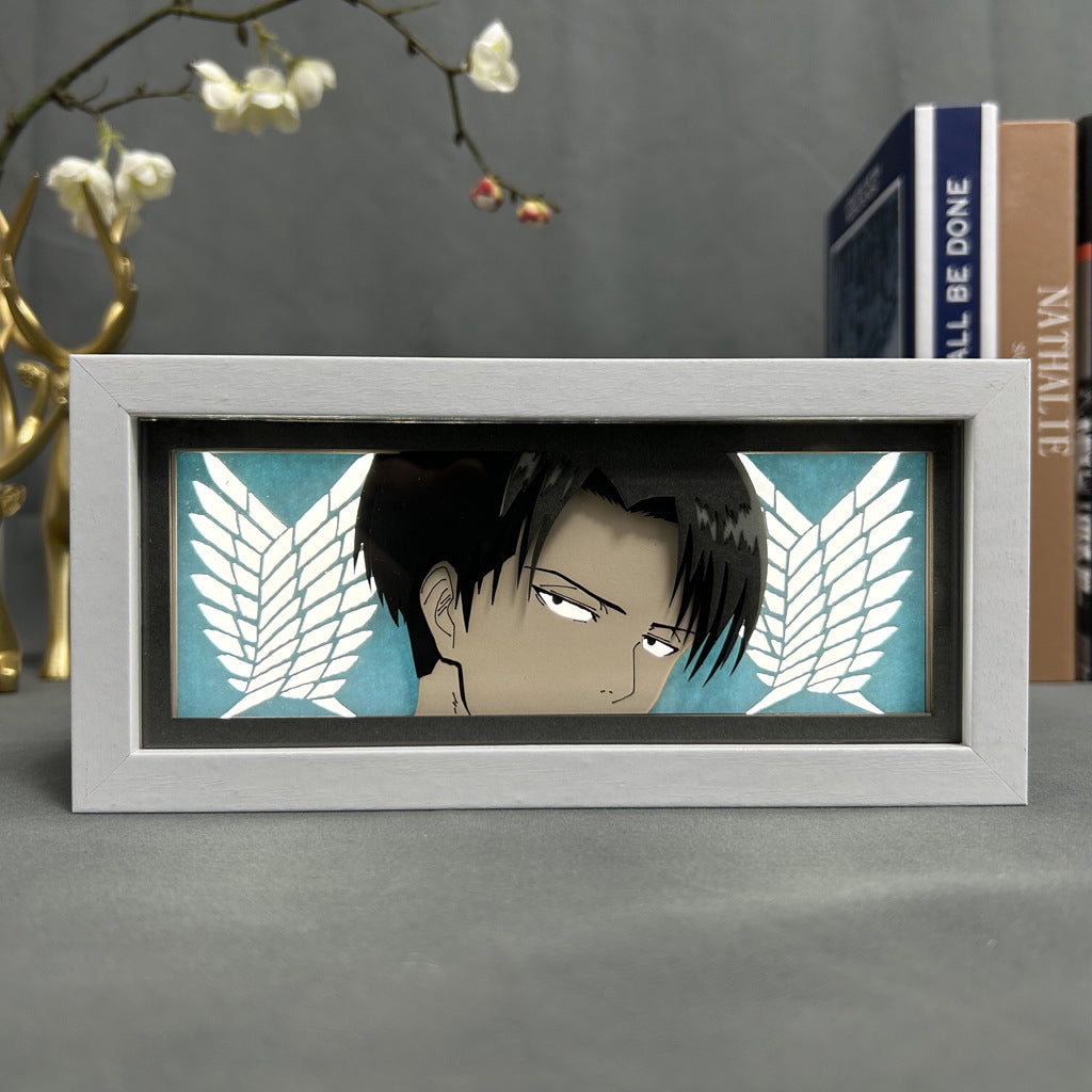 The Attack on Titan Anime Light Box features Levi Ackerman with the Wings of Freedom on a teal background. It includes a 3D effect and adjustable LED light in 16 colors. The box rests on a gray surface, with blurred plants and books behind it.