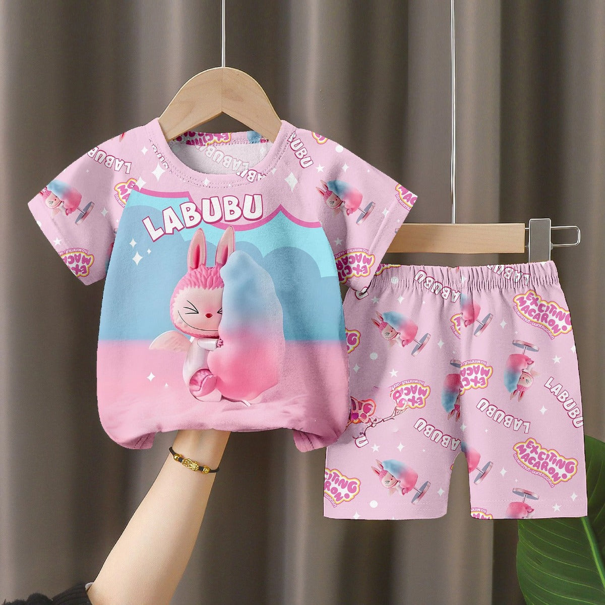 The LABUBU Baby Pajama Set, &quot;Exciting Macaron,&quot; features a pink, milk silk short-sleeved top and matching shorts. Displayed on a wooden hanger, it showcases a cartoon character with stars and &quot;LABUBU&quot; text against a gray curtain backdrop.