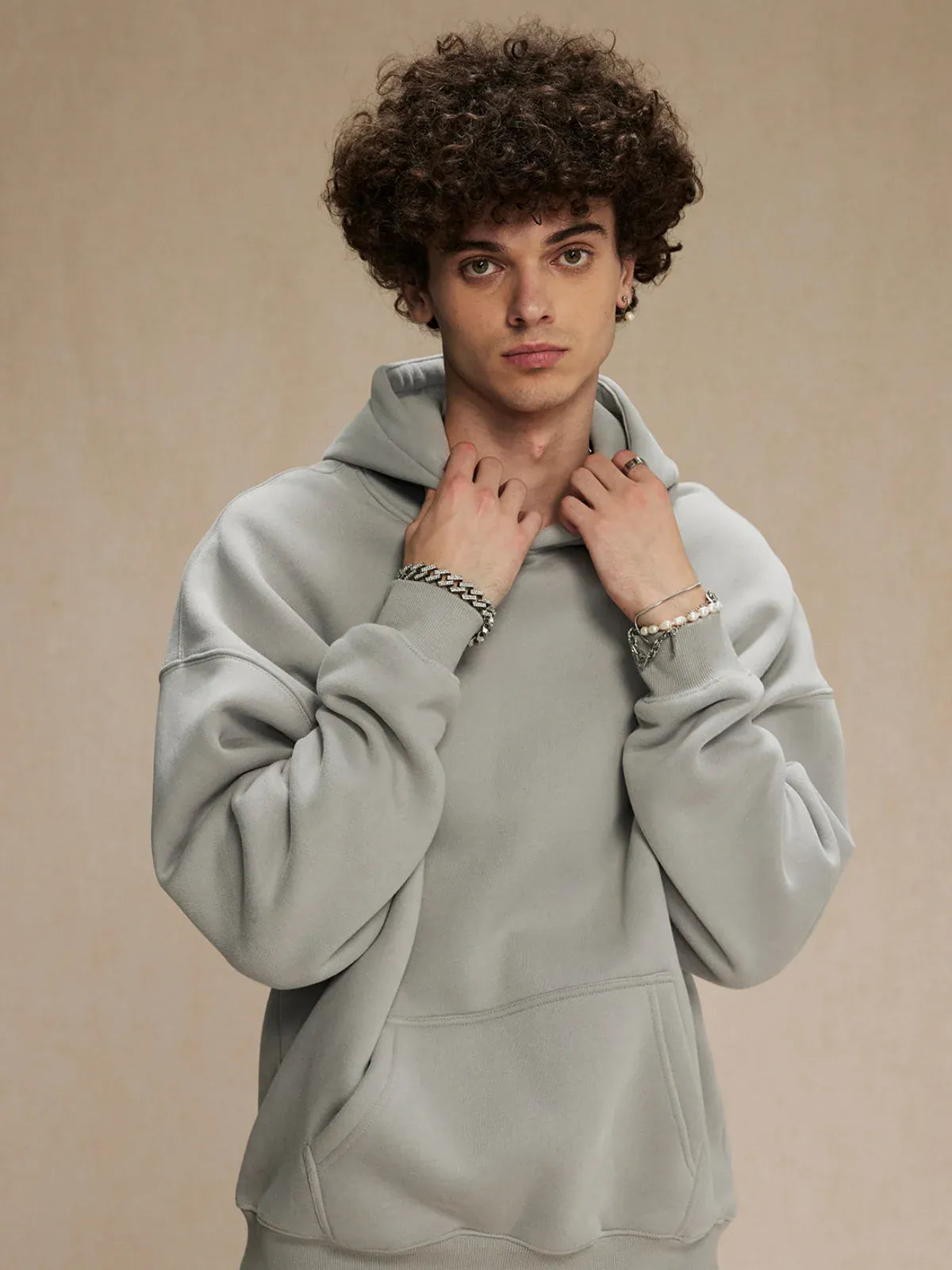 Young man with curly hair wears a Custom Anime Character Light Gray Hoodie—Personalized 350GSM 100% Cotton Pullover—plus bracelets and a watch, standing against a plain beige background.