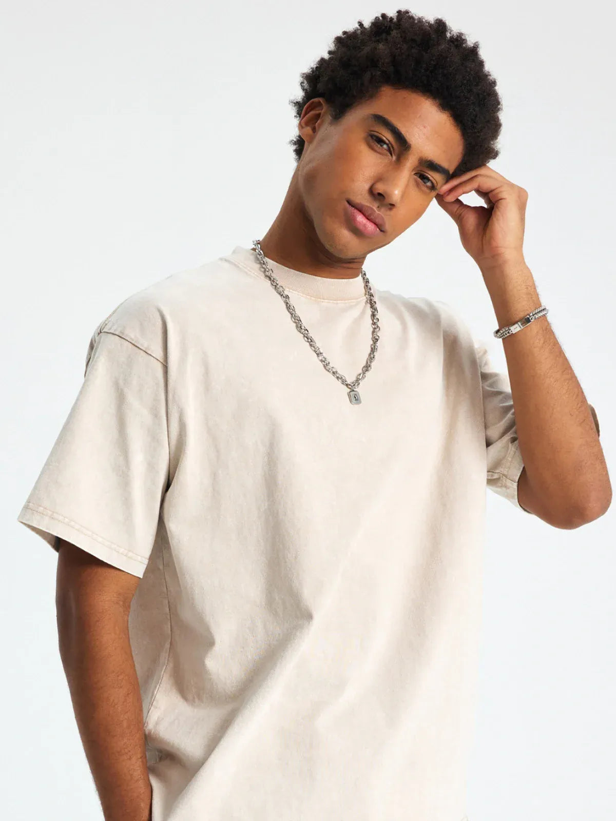 A man wearing a Custom Anime Character Beige Washed T-Shirt - Personalized Anime Style Tee, along with a chain necklace and bracelet, stands against a plain light background, touching his head with one hand.