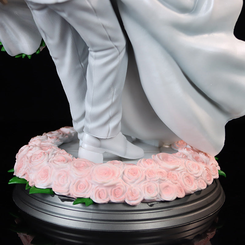 Close-up of the Seakoff Goku and Chi-Chi Wedding Collectible Figure – Dragon Ball Z. The bride and groom, inspired by Goku and Chi-Chi in elegant white attire, stand on a circular base with pink roses. Her dress flows gracefully while he sports a tuxedo. The base is dark-colored.