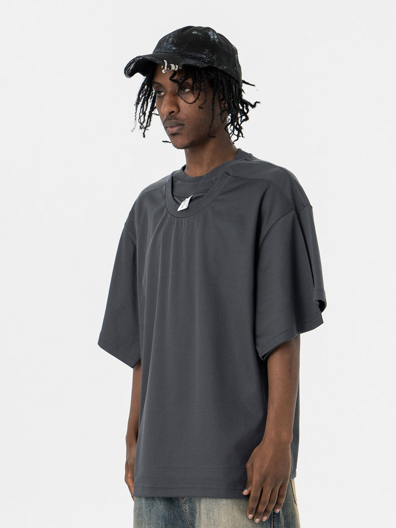 Oversized dark gray T-shirt with layered optical illusion neckline, worn by a model in a minimalist studio shot