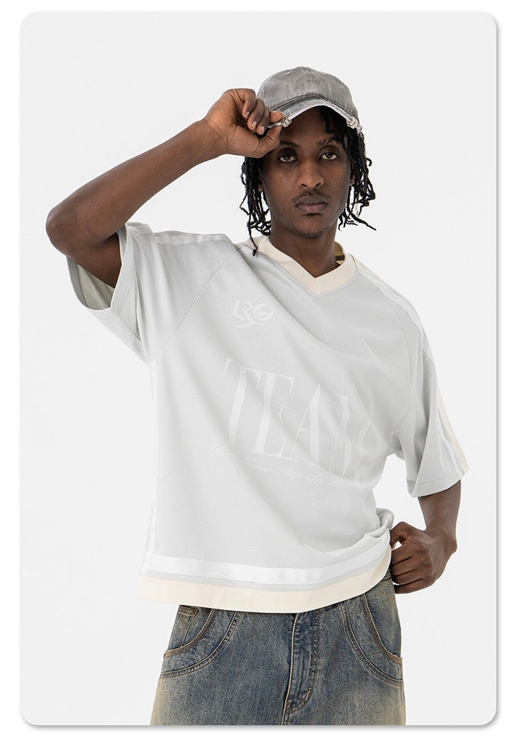 Model wearing an oversized white mesh varsity tee with faded lettering, paired with baggy jeans and a gray cap