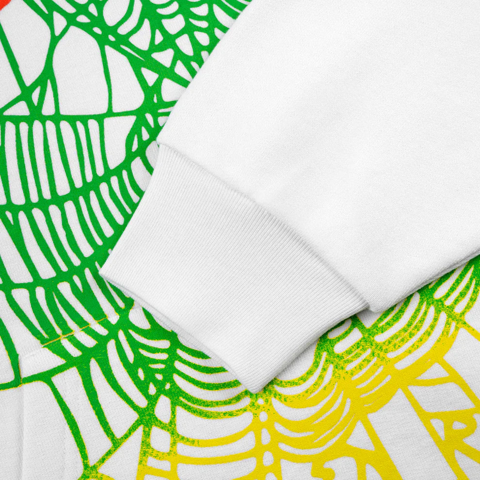 A close-up of a Sp5der “Toxic Web” Hoodie – White & Neon Lime, 440GSM heavyweight oversized streetwear pullover sleeve resting on fabric with an abstract green and yellow web-like pattern.